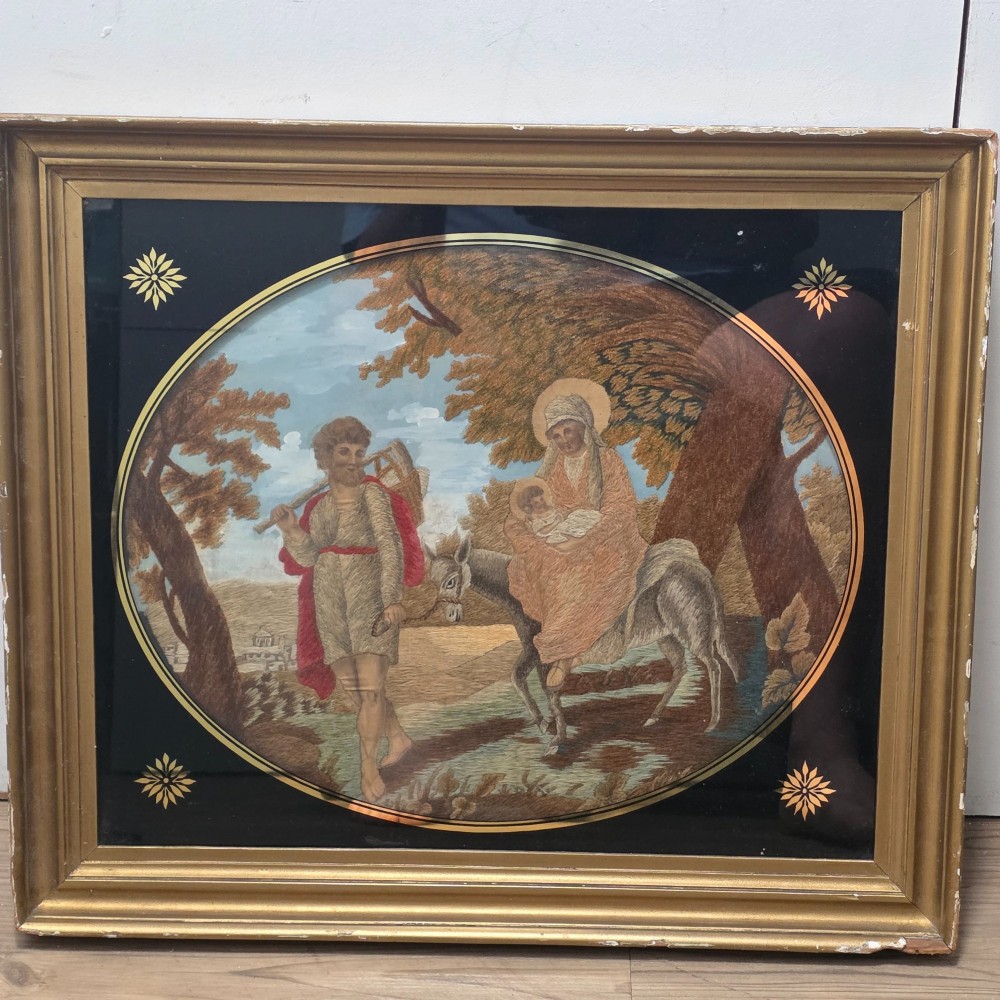 A framed Georgian silkwork and woolwork picture depicting the Flight into Egypt inside an oval glass reserve and a giltwood frame.