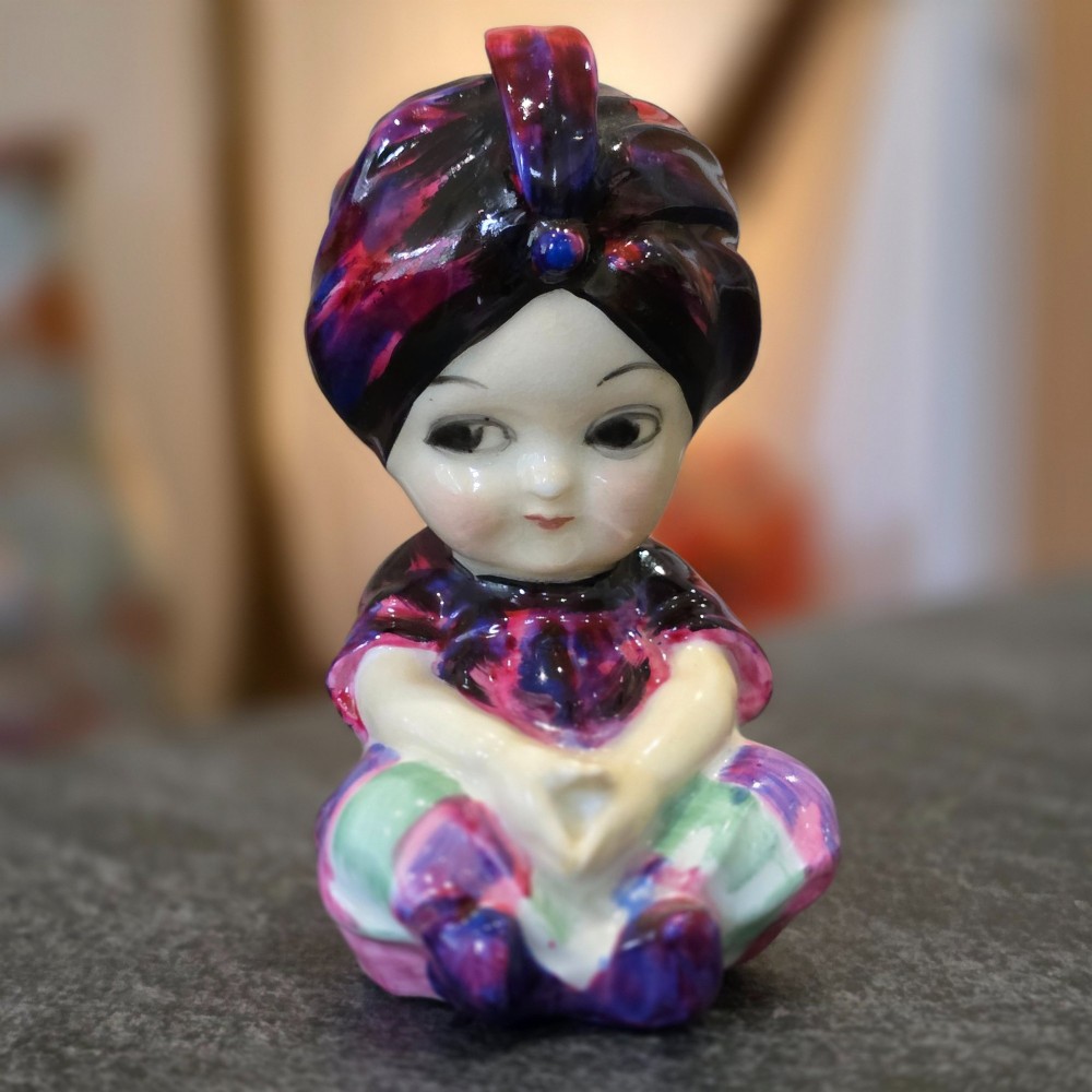 A small glazed ceramic figurine depicting a seated child wearing a patterned purple turban.