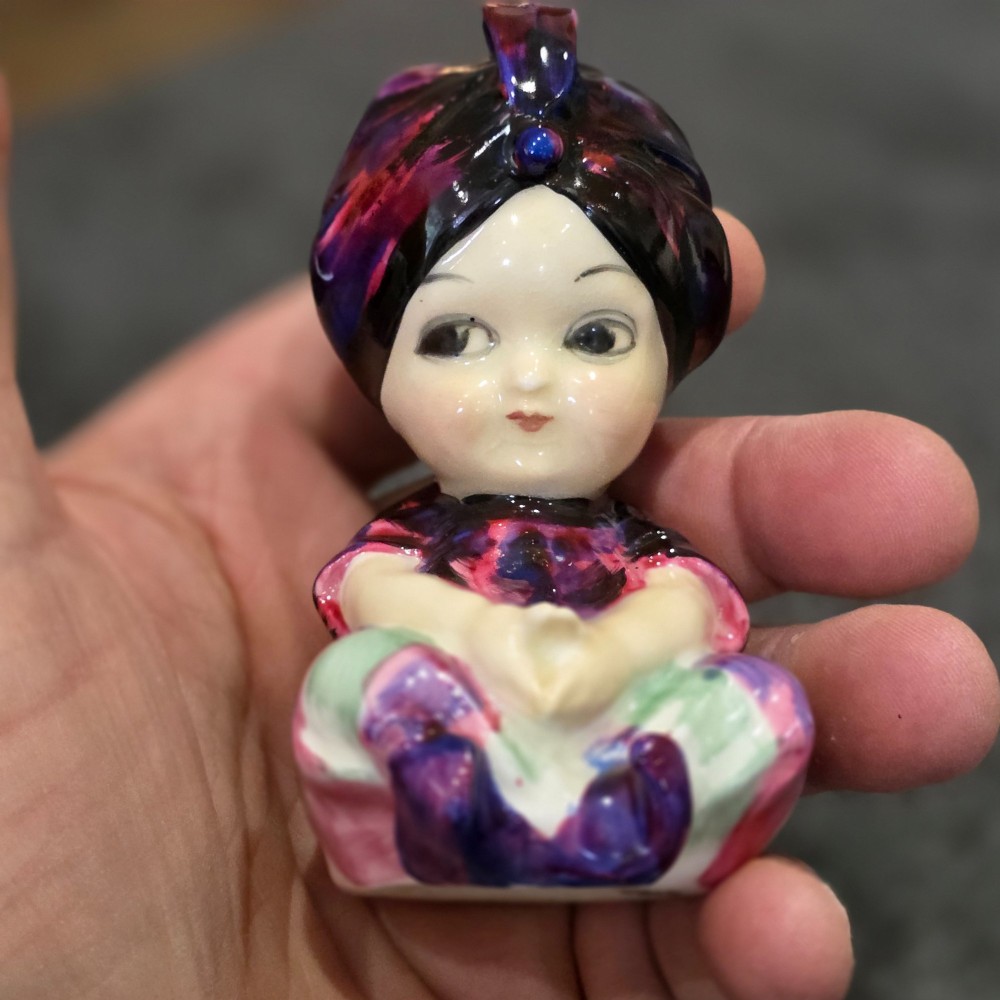 A small glazed ceramic figurine depicting a seated child wearing a patterned purple turban. View II