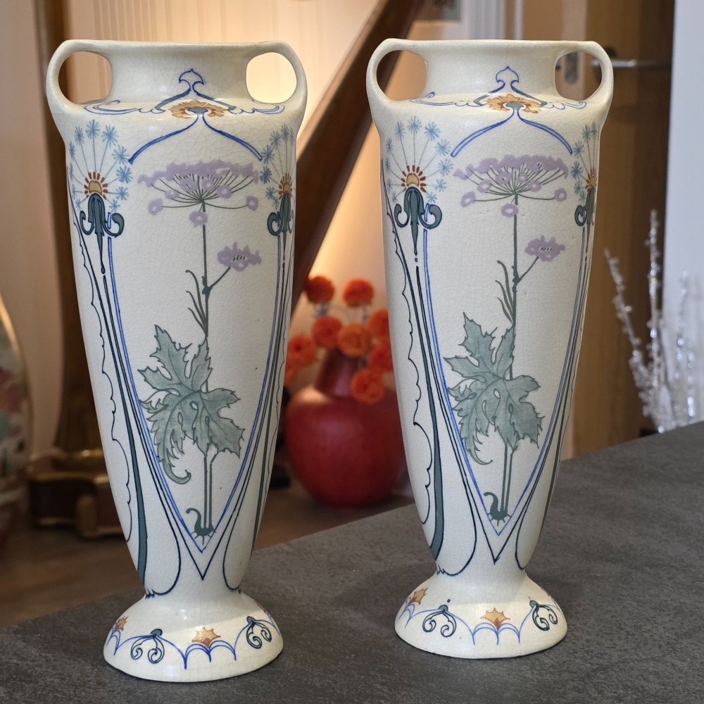 A pair of tall cream ceramic Art Nouveau vases featuring integral twin handles and hand-painted purple and green stylised floral motifs.