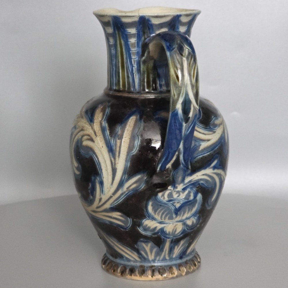 A Victorian salt-glazed stoneware jug decorated with incised blue and cream foliage patterns, a ribbed neck, and a loop handle against a dark brown ground. View V