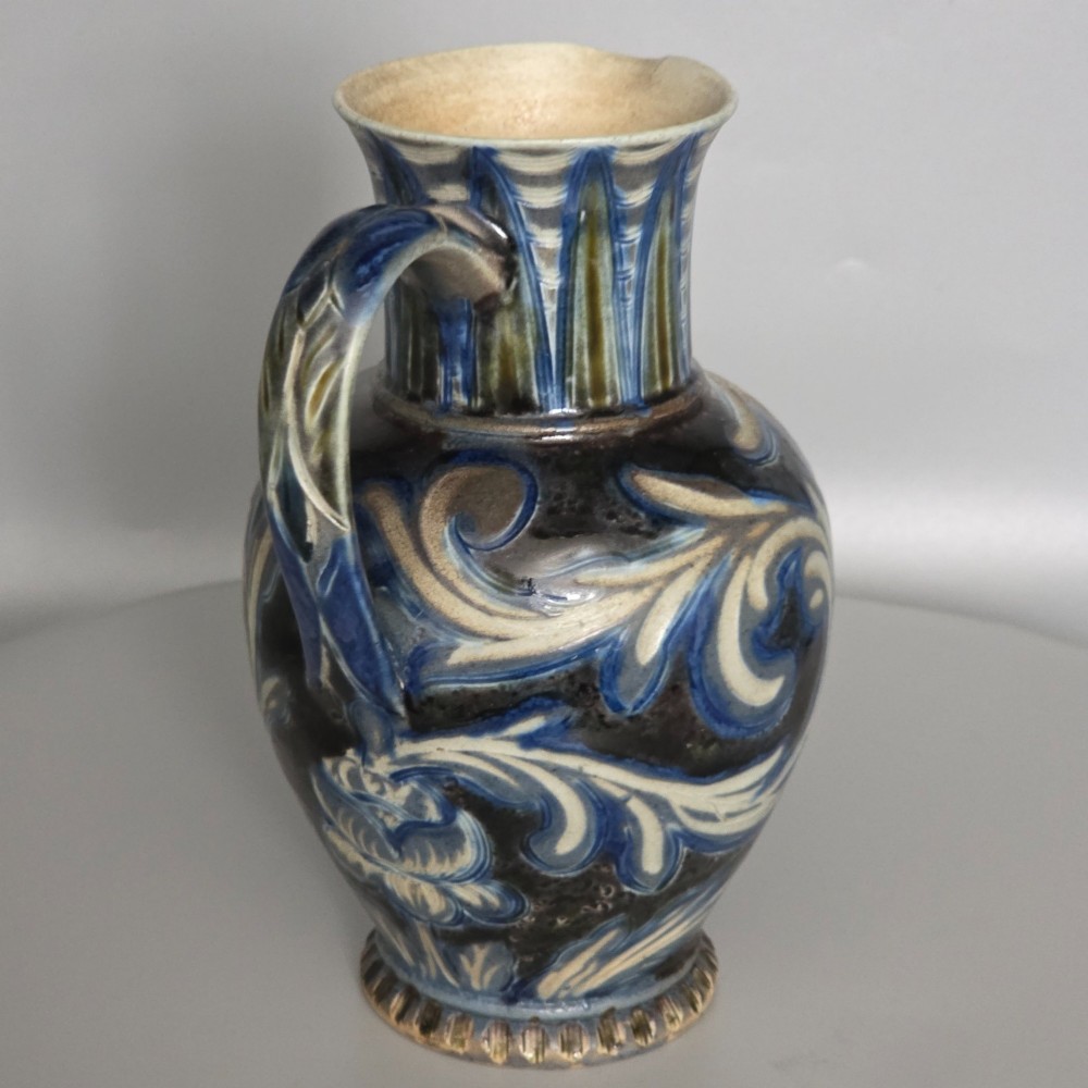 A Victorian salt-glazed stoneware jug decorated with incised blue and cream foliage patterns, a ribbed neck, and a loop handle against a dark brown ground. View VI
