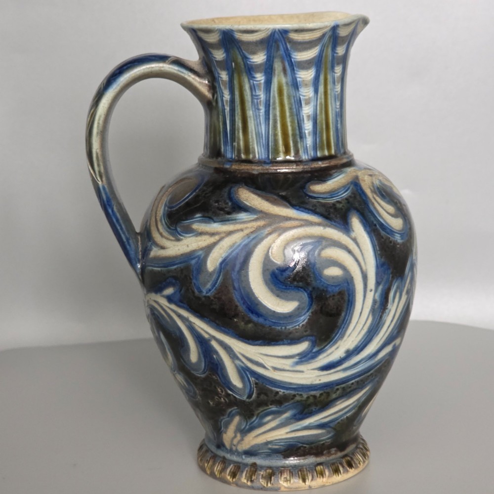 A Victorian salt-glazed stoneware jug decorated with incised blue and cream foliage patterns, a ribbed neck, and a loop handle against a dark brown ground. View VII