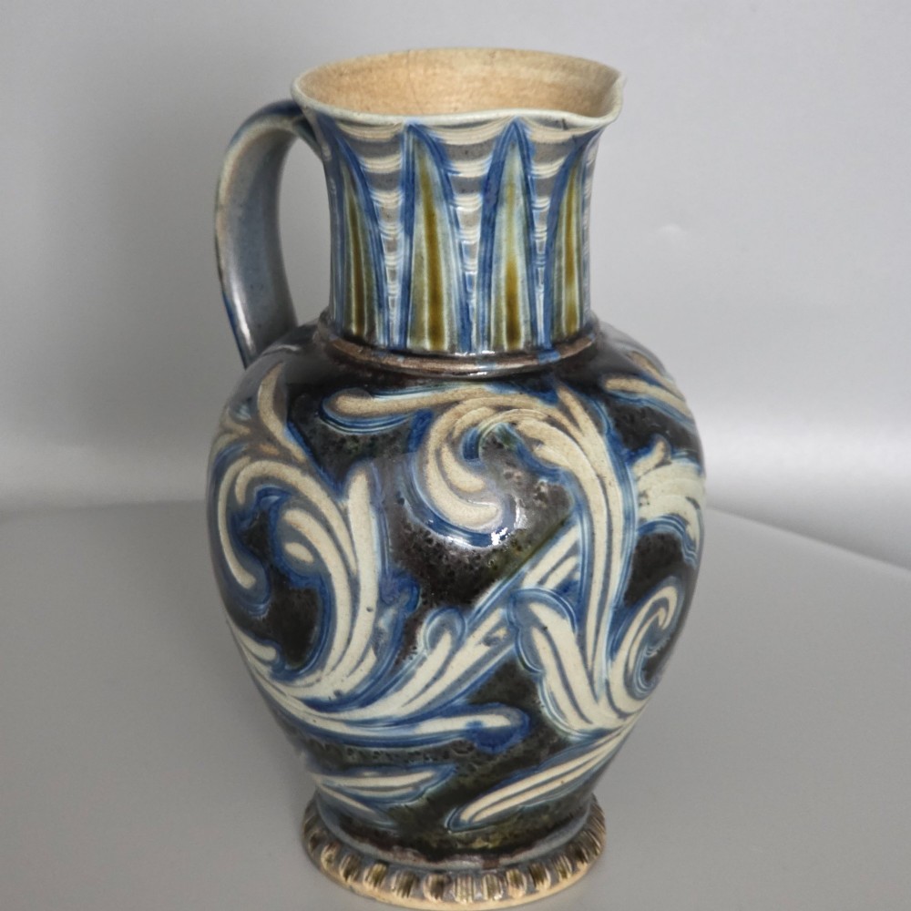 A Victorian salt-glazed stoneware jug decorated with incised blue and cream foliage patterns, a ribbed neck, and a loop handle against a dark brown ground. View VIII
