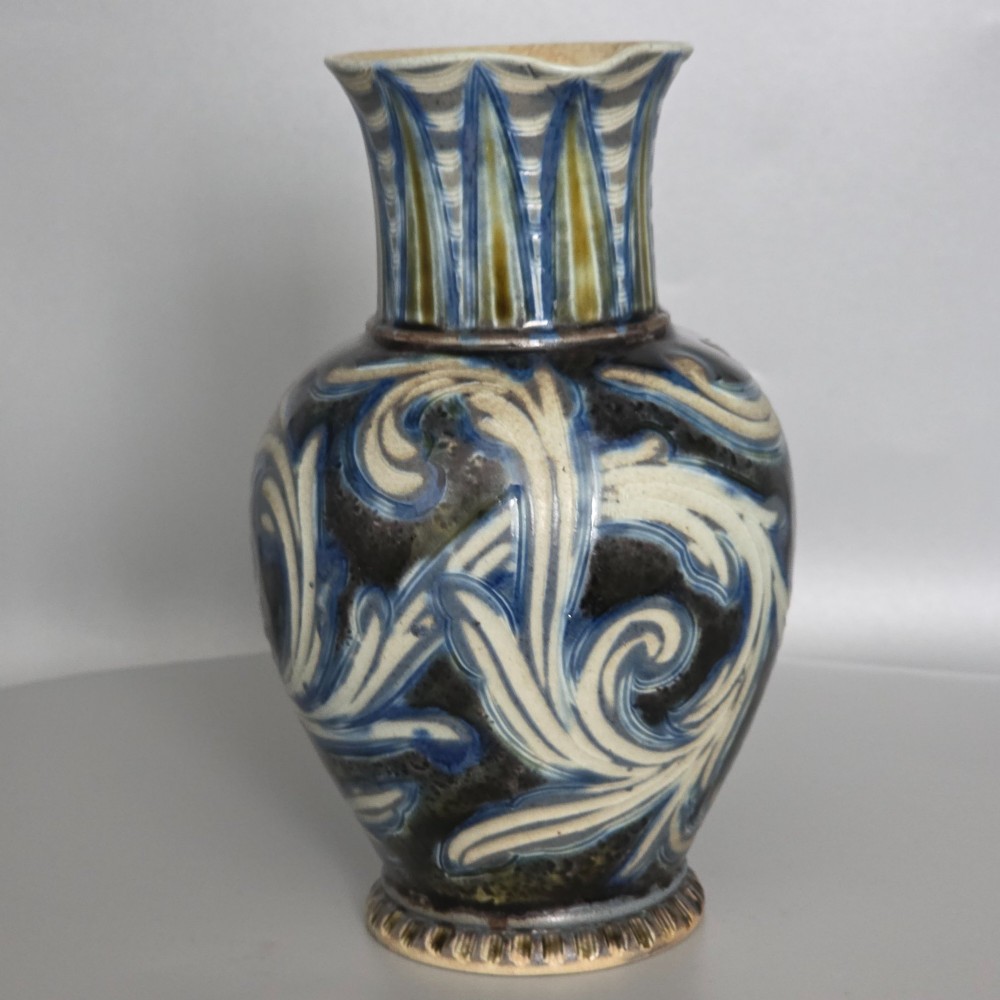 A Victorian salt-glazed stoneware jug decorated with incised blue and cream foliage patterns, a ribbed neck, and a loop handle against a dark brown ground. View IX
