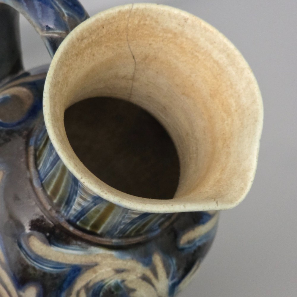A Victorian salt-glazed stoneware jug decorated with incised blue and cream foliage patterns, a ribbed neck, and a loop handle against a dark brown ground. View X