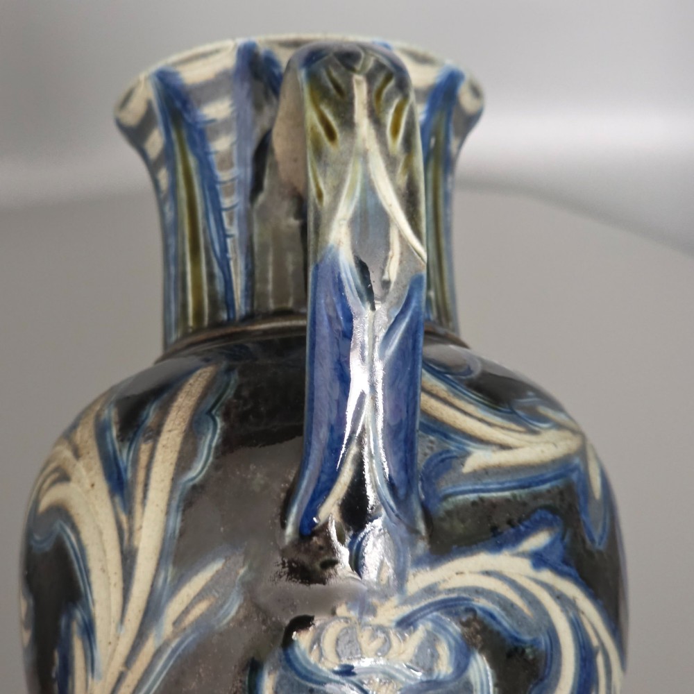 A Victorian salt-glazed stoneware jug decorated with incised blue and cream foliage patterns, a ribbed neck, and a loop handle against a dark brown ground. View XIII