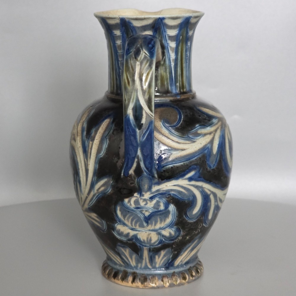 A Victorian salt-glazed stoneware jug decorated with incised blue and cream foliage patterns, a ribbed neck, and a loop handle against a dark brown ground. View XIV