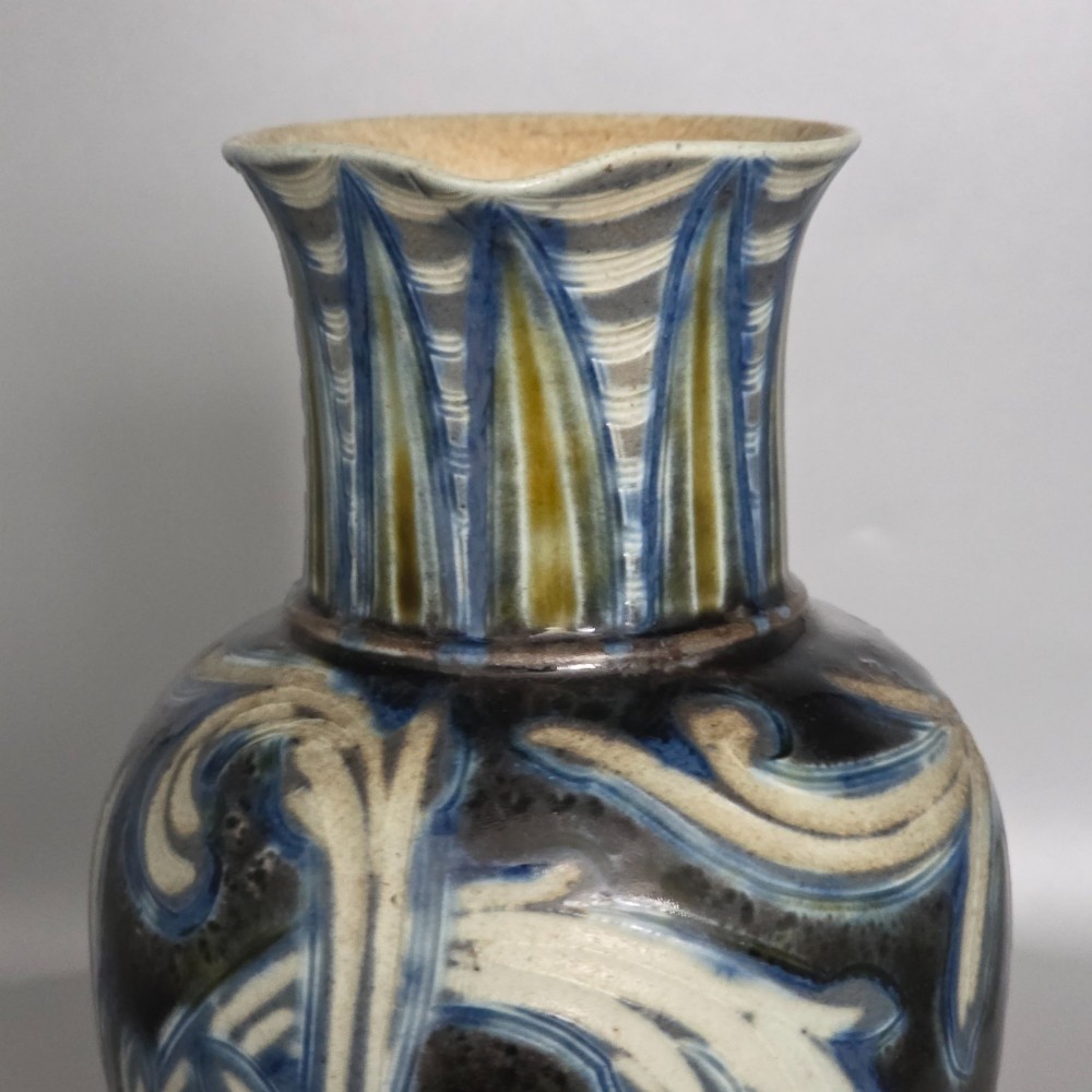 A Victorian salt-glazed stoneware jug decorated with incised blue and cream foliage patterns, a ribbed neck, and a loop handle against a dark brown ground. View XV