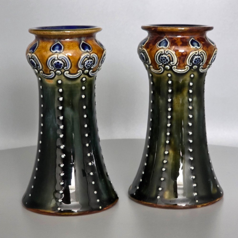 A pair of 1920s Royal Doulton Art Nouveau stoneware vases featuring flared bases, mottled dark green and brown glazes, and beaded shoulder decorations with blue accents. View X