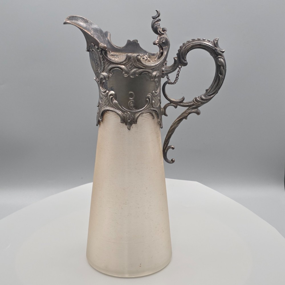 An antique French Rococo style silver-plated claret jug featuring a tapered ribbed clear glass body and an ornate scrolling silver spout and handle. View II