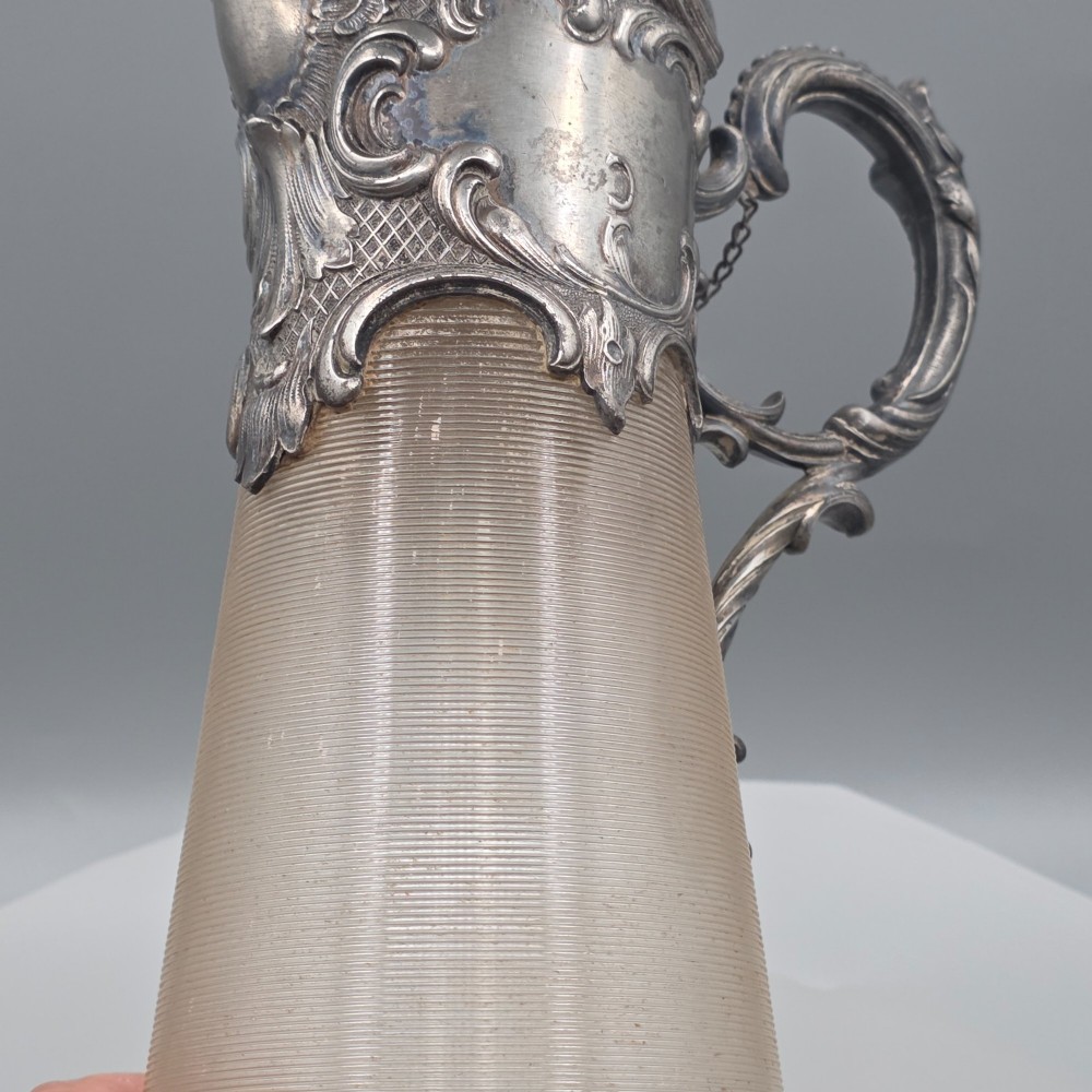 An antique French Rococo style silver-plated claret jug featuring a tapered ribbed clear glass body and an ornate scrolling silver spout and handle. View XV