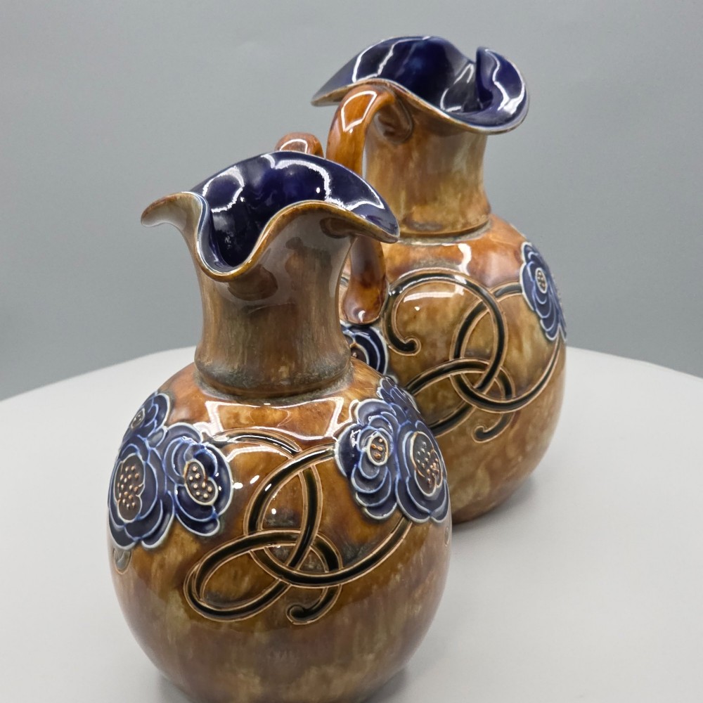 A pair of Royal Doulton Arts and Crafts glazed stoneware jugs featuring mottled brown bodies, deep blue interiors, and stylised tube-lined floral motifs. View II