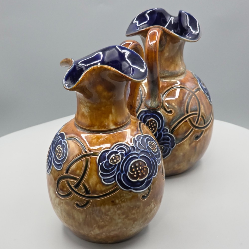 A pair of Royal Doulton Arts and Crafts glazed stoneware jugs featuring mottled brown bodies, deep blue interiors, and stylised tube-lined floral motifs. View V