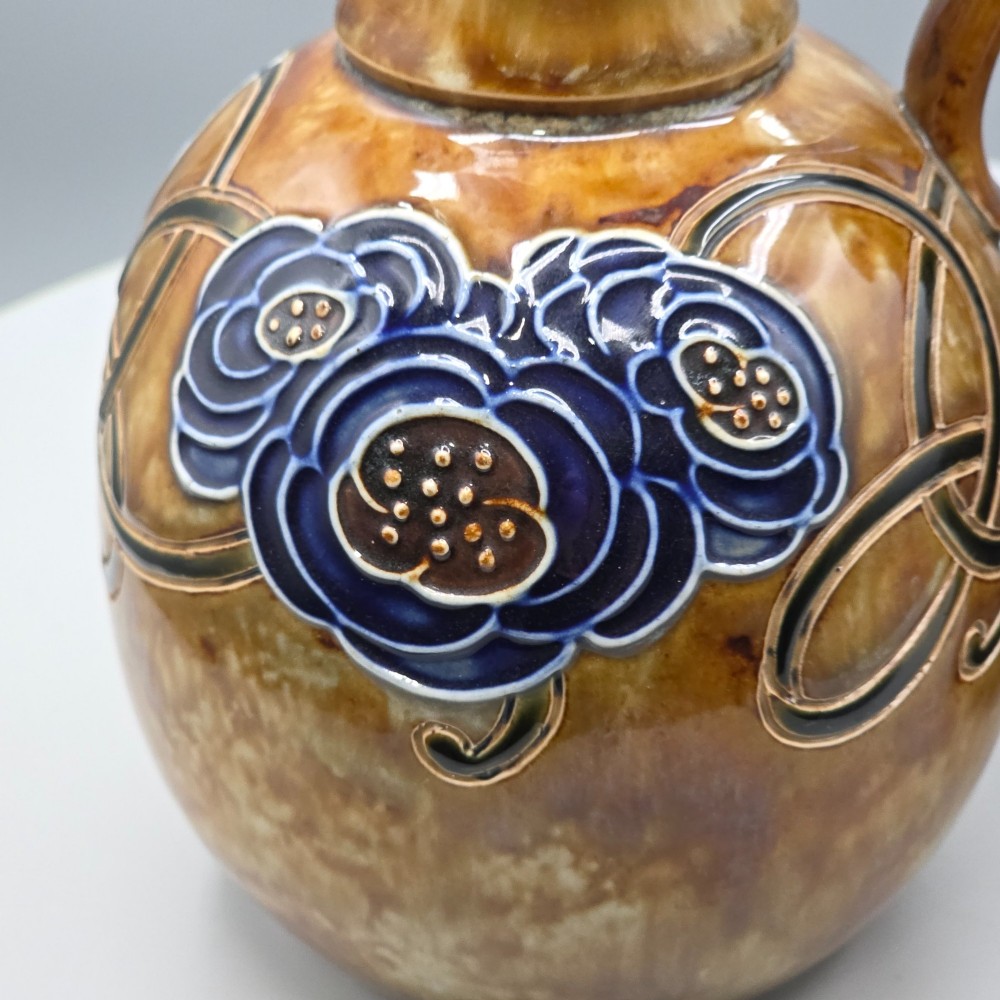A pair of Royal Doulton Arts and Crafts glazed stoneware jugs featuring mottled brown bodies, deep blue interiors, and stylised tube-lined floral motifs. View VI