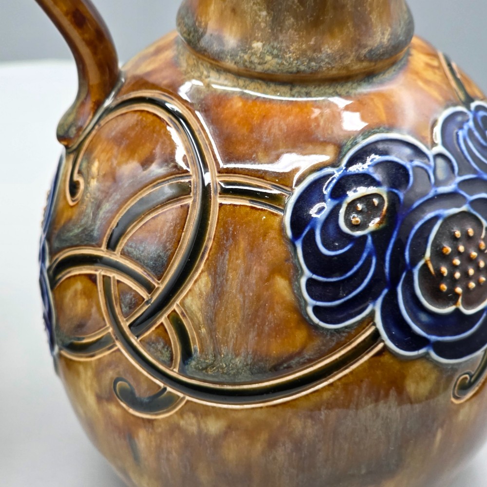 A pair of Royal Doulton Arts and Crafts glazed stoneware jugs featuring mottled brown bodies, deep blue interiors, and stylised tube-lined floral motifs. View VII