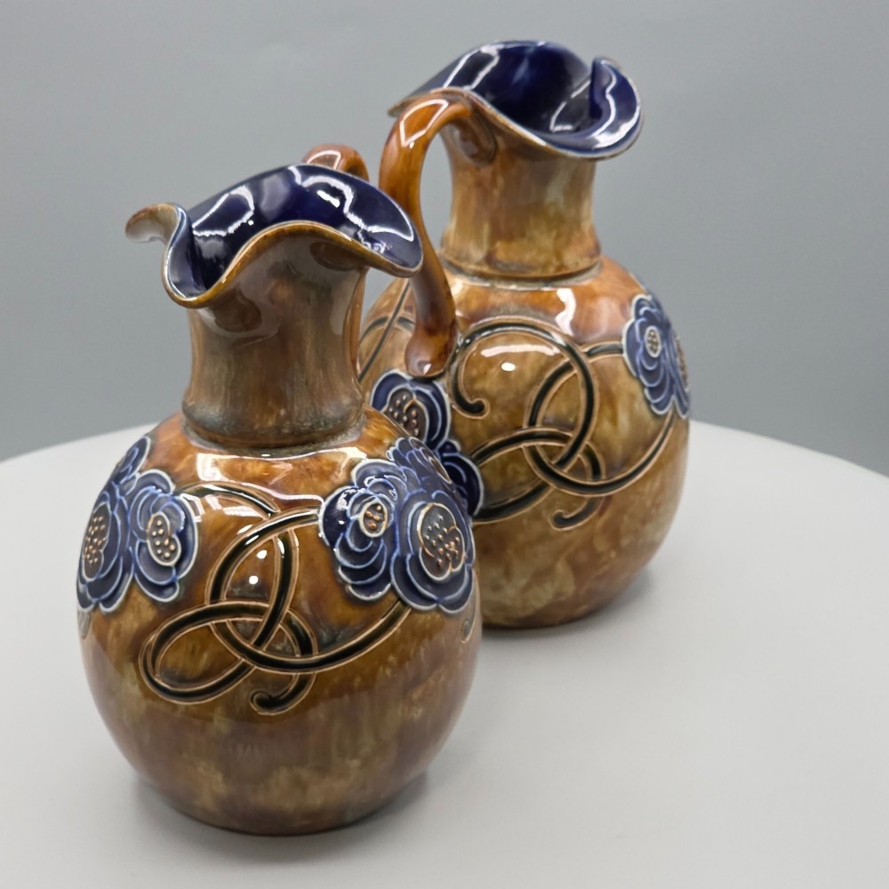 A pair of Royal Doulton Arts and Crafts glazed stoneware jugs featuring mottled brown bodies, deep blue interiors, and stylised tube-lined floral motifs. View VIII