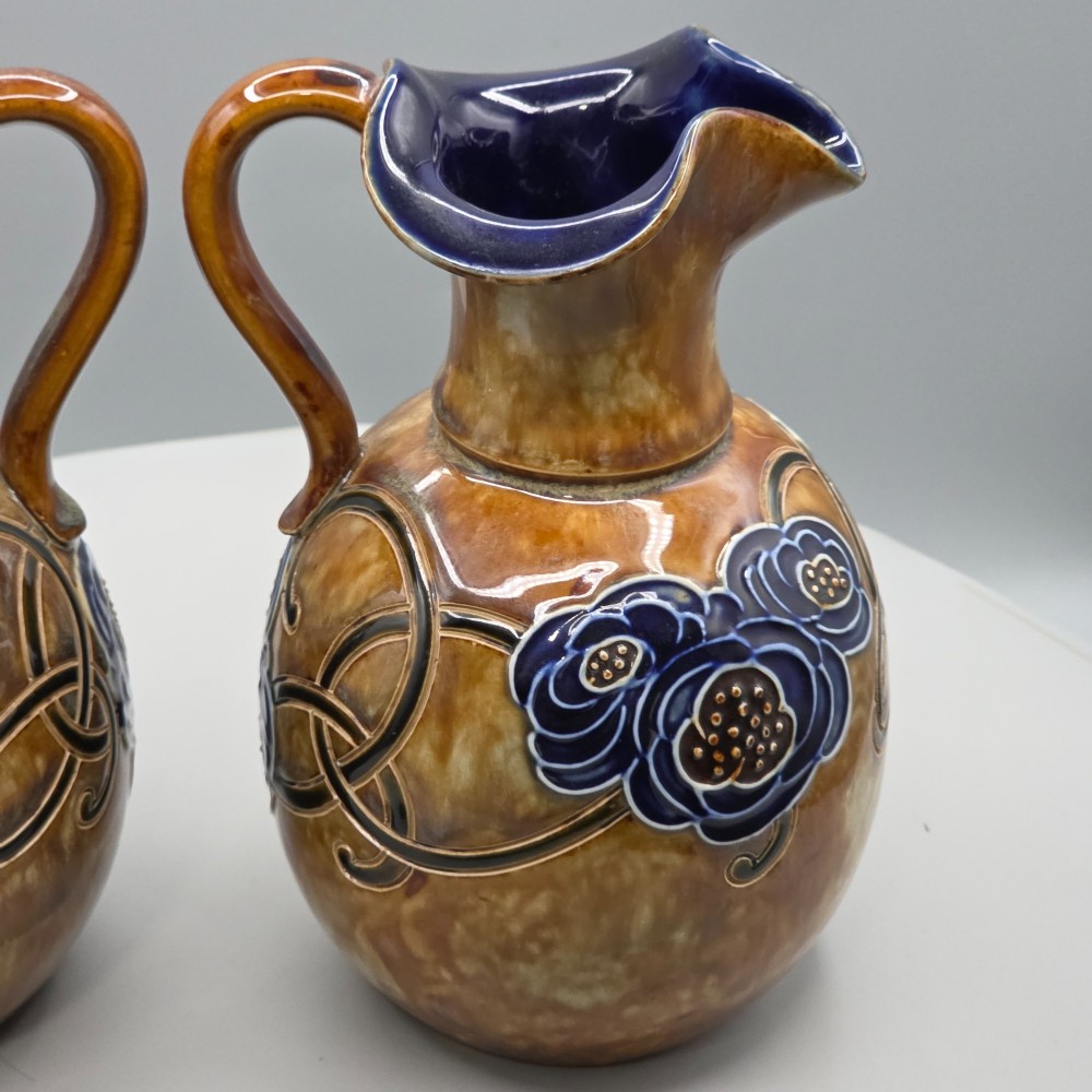 A pair of Royal Doulton Arts and Crafts glazed stoneware jugs featuring mottled brown bodies, deep blue interiors, and stylised tube-lined floral motifs. View IX