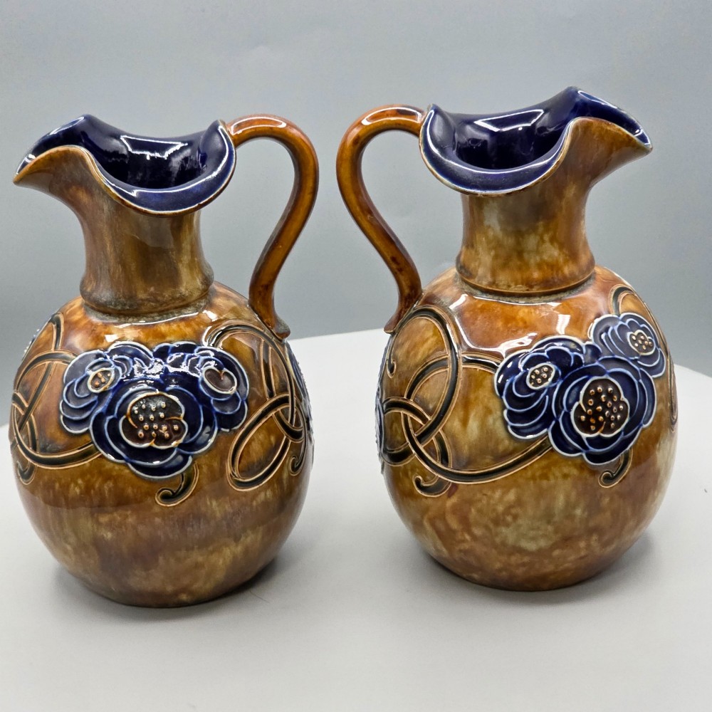 A pair of Royal Doulton Arts and Crafts glazed stoneware jugs featuring mottled brown bodies, deep blue interiors, and stylised tube-lined floral motifs. View X