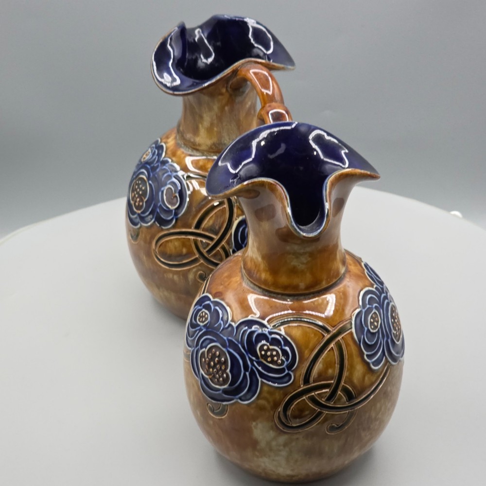 A pair of Royal Doulton Arts and Crafts glazed stoneware jugs featuring mottled brown bodies, deep blue interiors, and stylised tube-lined floral motifs. View XI
