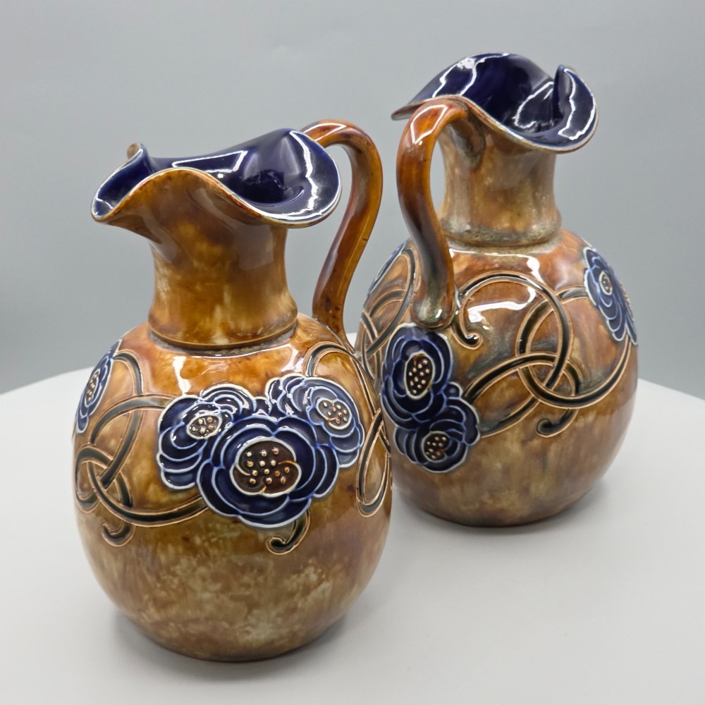 A pair of Royal Doulton Arts and Crafts glazed stoneware jugs featuring mottled brown bodies, deep blue interiors, and stylised tube-lined floral motifs. View XII