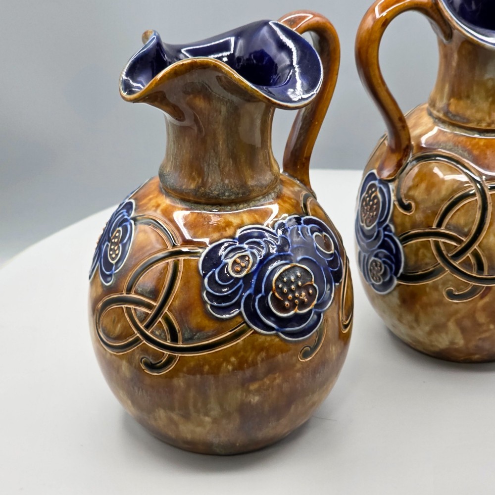 A pair of Royal Doulton Arts and Crafts glazed stoneware jugs featuring mottled brown bodies, deep blue interiors, and stylised tube-lined floral motifs. View XIII