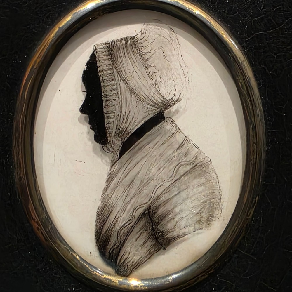 An antique oval Georgian silhouette portrait depicting a woman in profile wearing a bonnet, rendered via reverse painting on glass within a gilded bezel.