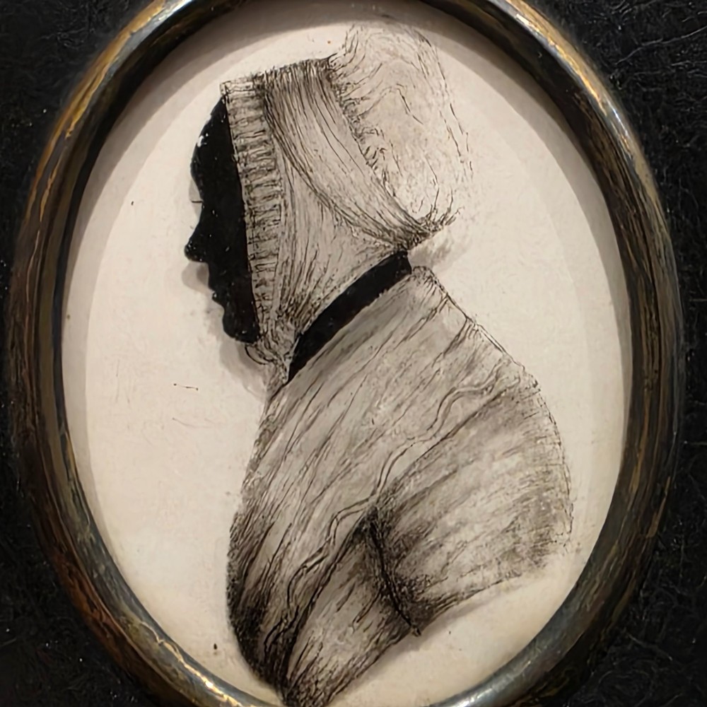 An antique oval Georgian silhouette portrait depicting a woman in profile wearing a bonnet, rendered via reverse painting on glass within a gilded bezel. View II
