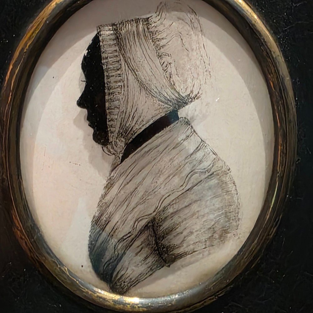 An antique oval Georgian silhouette portrait depicting a woman in profile wearing a bonnet, rendered via reverse painting on glass within a gilded bezel. View III