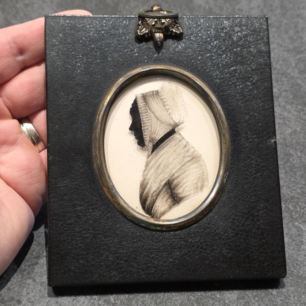 An antique oval Georgian silhouette portrait depicting a woman in profile wearing a bonnet, rendered via reverse painting on glass within a gilded bezel. View V