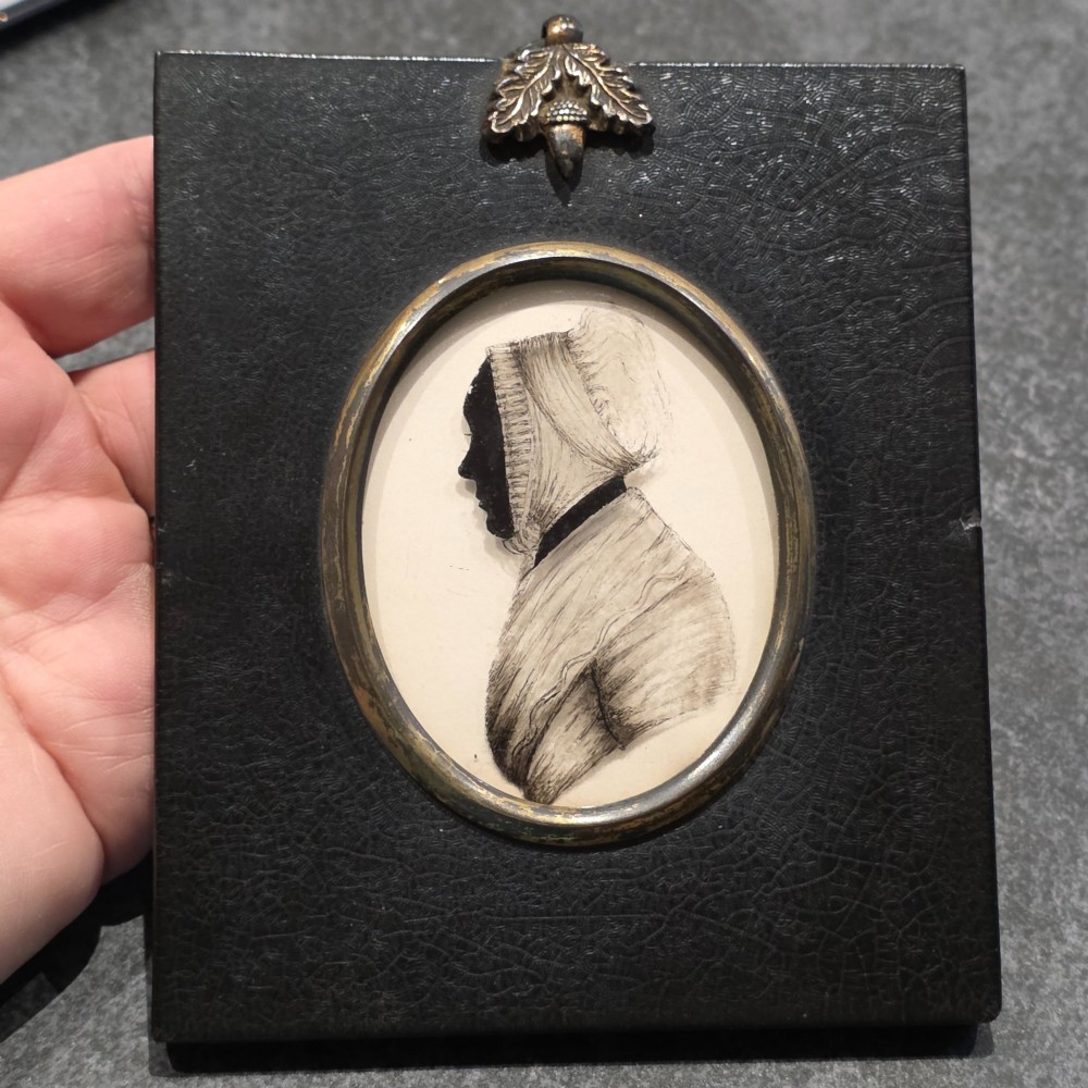 An antique oval Georgian silhouette portrait depicting a woman in profile wearing a bonnet, rendered via reverse painting on glass within a gilded bezel. View VIII