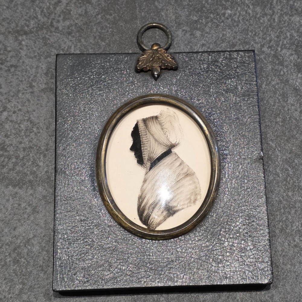 An antique oval Georgian silhouette portrait depicting a woman in profile wearing a bonnet, rendered via reverse painting on glass within a gilded bezel. View IX