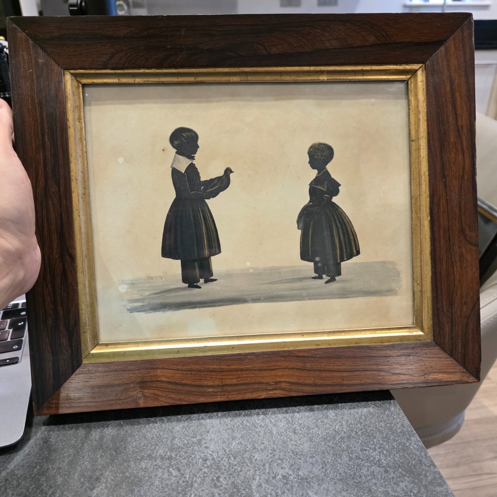 A framed 19th-century silhouette portrait depicting two standing children, one holding a bird, enclosed in a wide timber frame with a gilded inner slip.