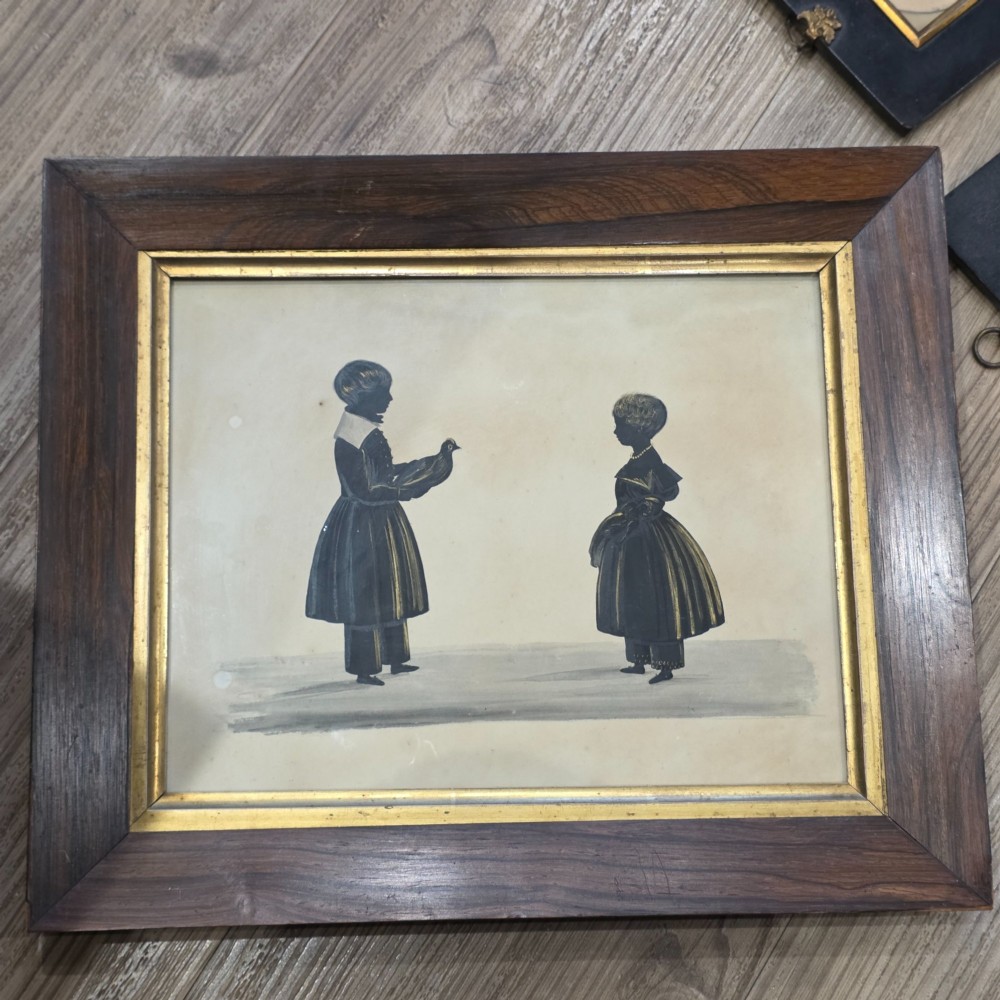 A framed 19th-century silhouette portrait depicting two standing children, one holding a bird, enclosed in a wide timber frame with a gilded inner slip. View II