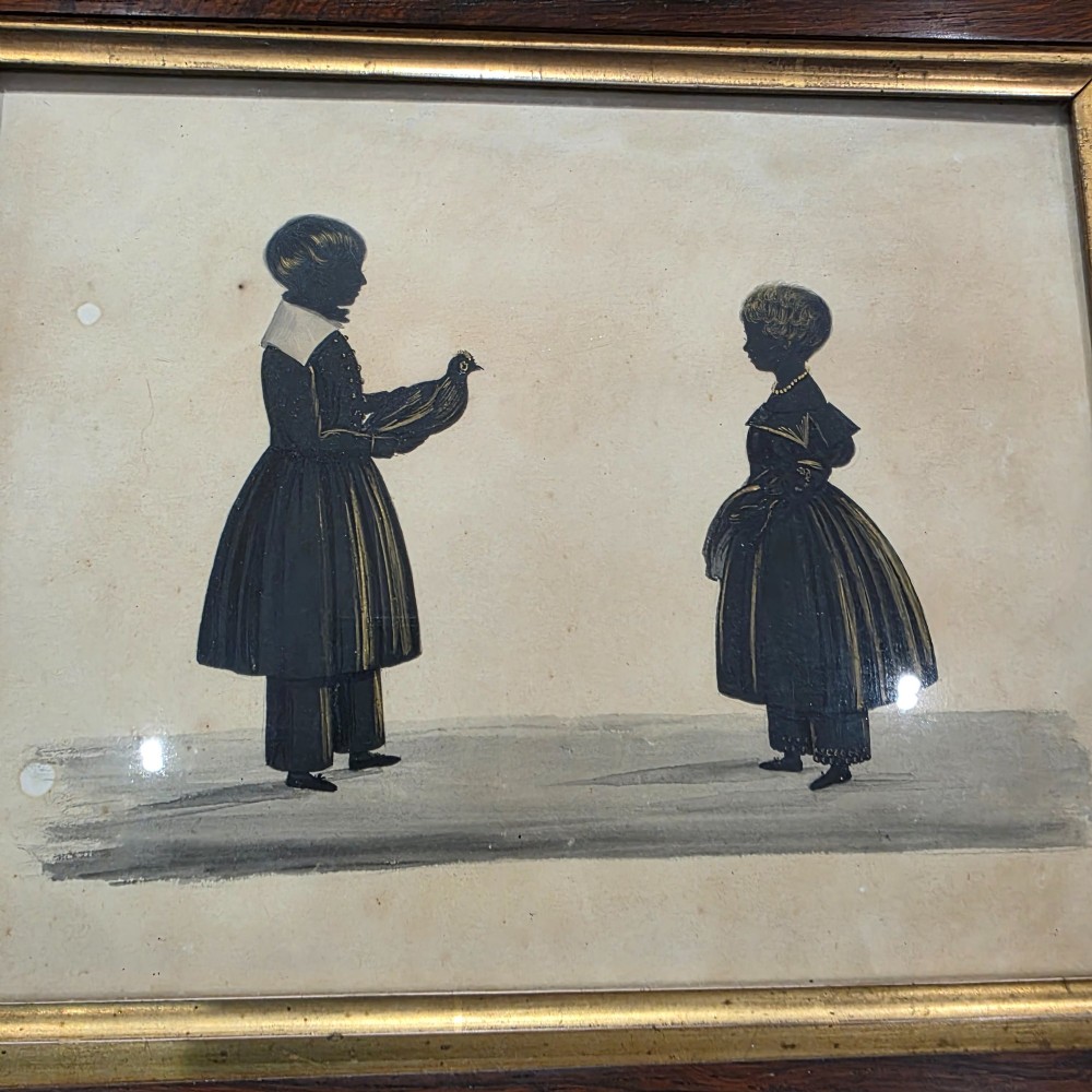 A framed 19th-century silhouette portrait depicting two standing children, one holding a bird, enclosed in a wide timber frame with a gilded inner slip. View III