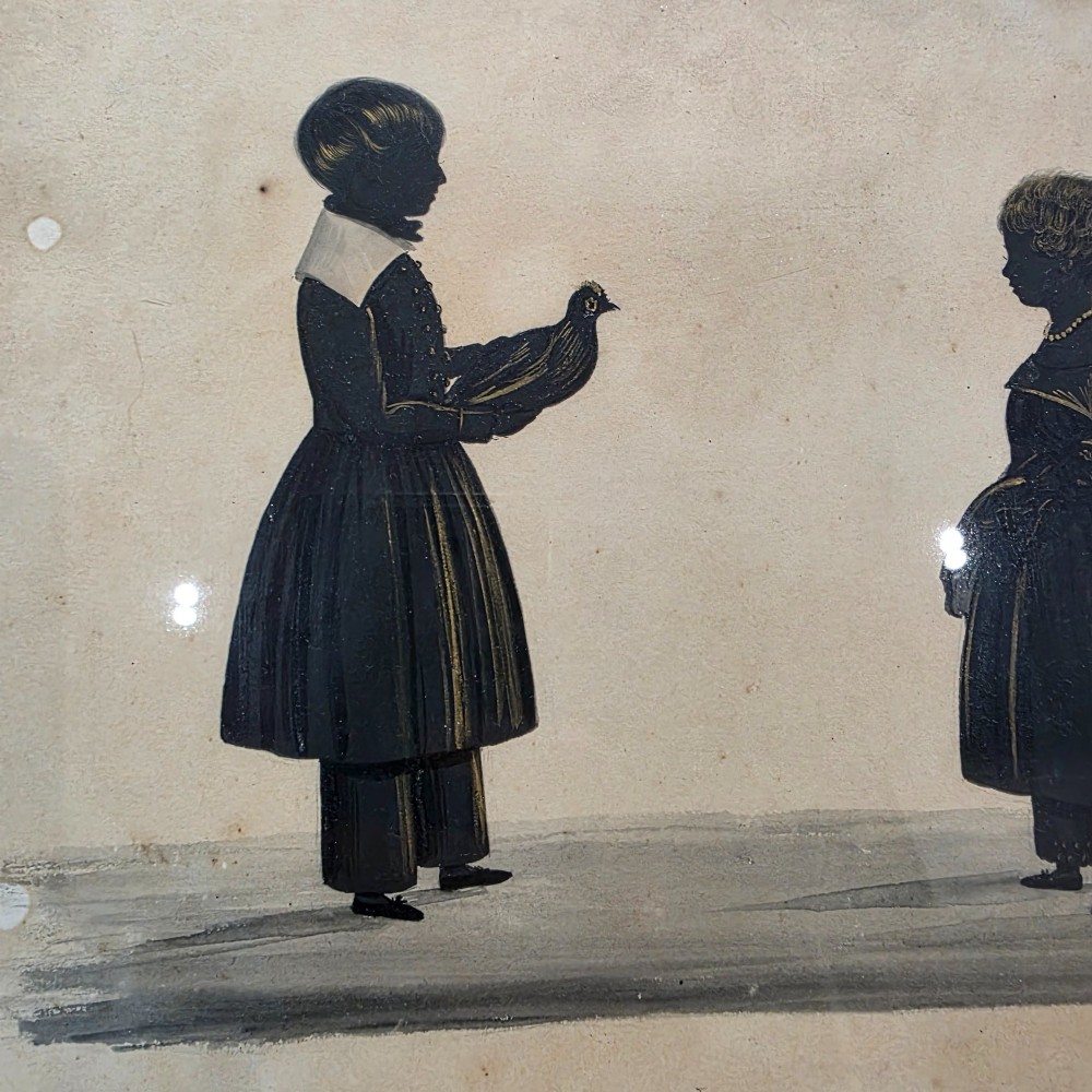 A framed 19th-century silhouette portrait depicting two standing children, one holding a bird, enclosed in a wide timber frame with a gilded inner slip. View IV