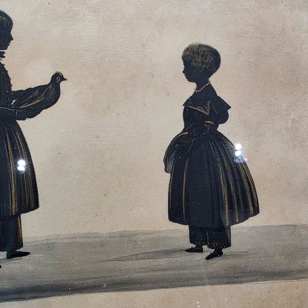 A framed 19th-century silhouette portrait depicting two standing children, one holding a bird, enclosed in a wide timber frame with a gilded inner slip. View V