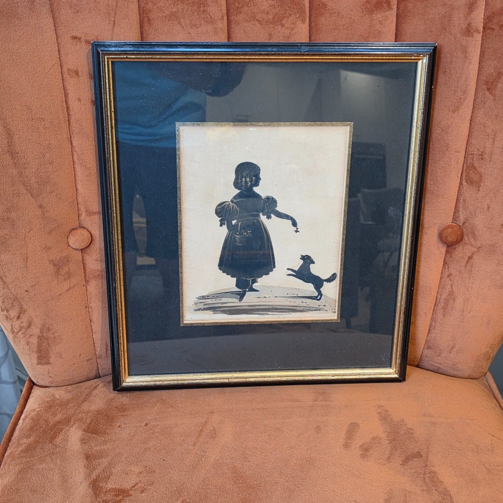 A framed antique cut paper silhouette depicting a young girl holding a basket alongside a small leaping dog.