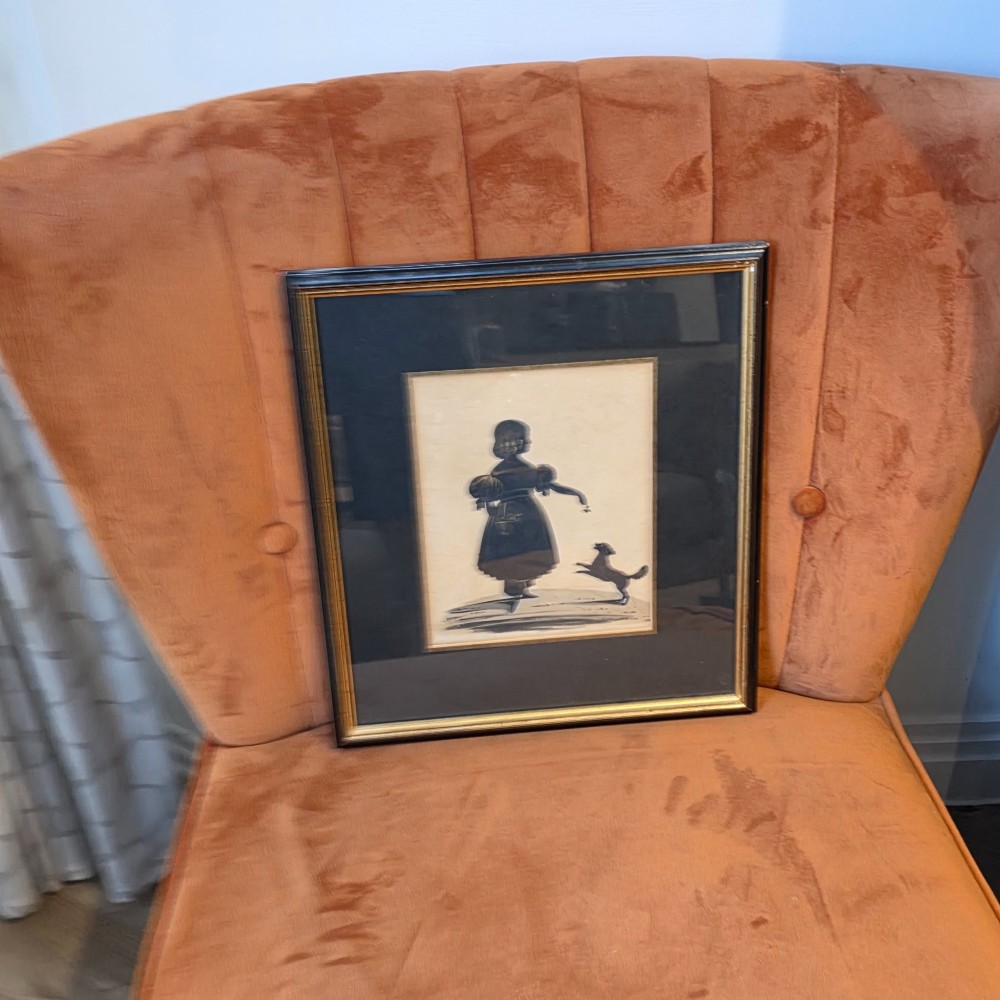 A framed antique cut paper silhouette depicting a young girl holding a basket alongside a small leaping dog. View II