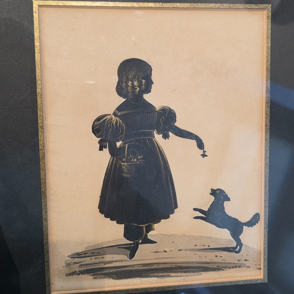 A framed antique cut paper silhouette depicting a young girl holding a basket alongside a small leaping dog. View III