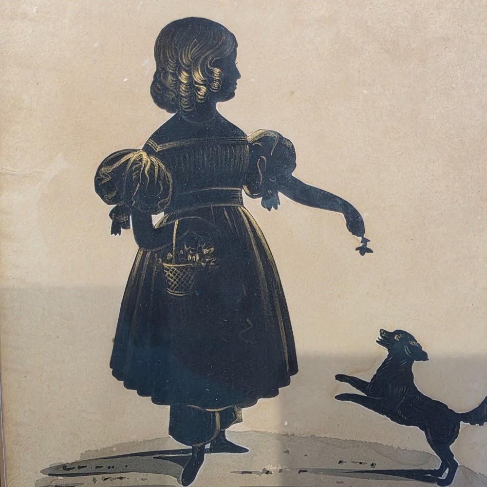 A framed antique cut paper silhouette depicting a young girl holding a basket alongside a small leaping dog. View IV