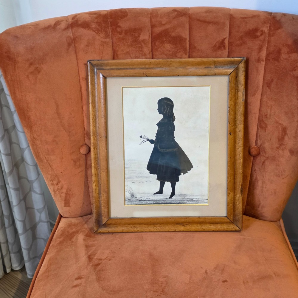 A 19th-century full-length cut-paper silhouette portrait of a young girl holding flowers, displayed in a glazed wooden frame with a gold inner slip. View II