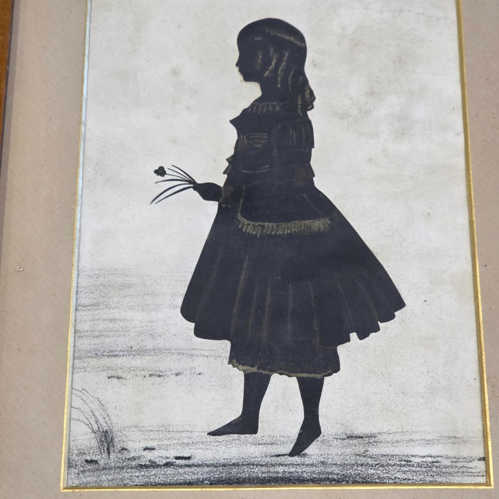 A 19th-century full-length cut-paper silhouette portrait of a young girl holding flowers, displayed in a glazed wooden frame with a gold inner slip. View IV