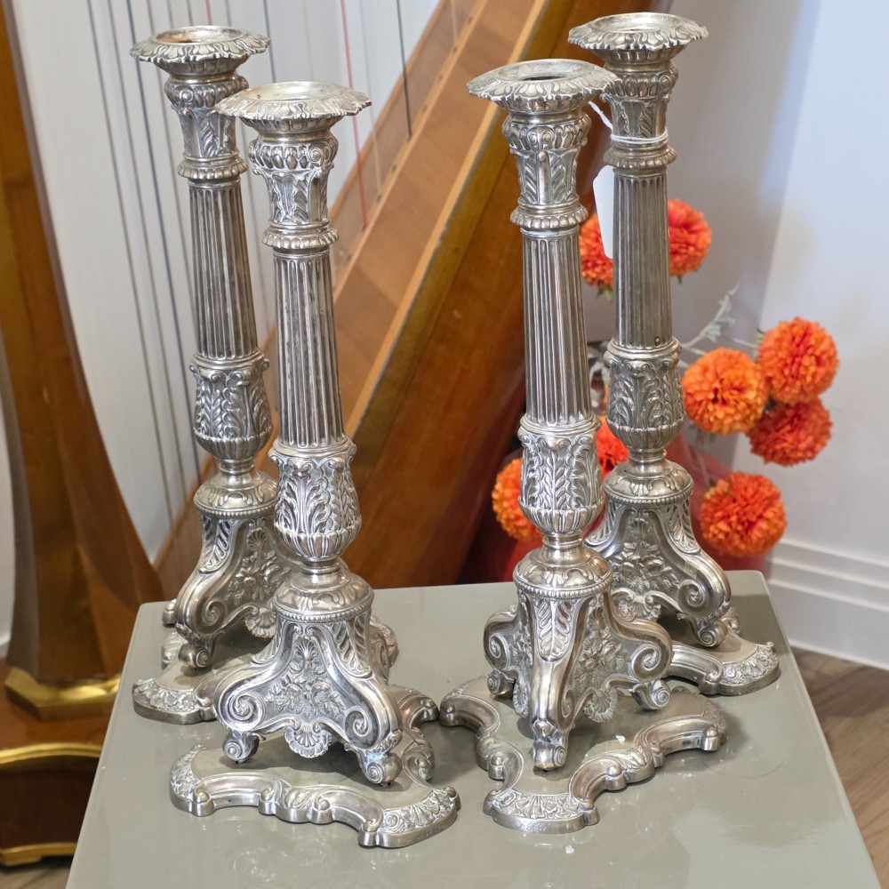 A set of four silver candlesticks with fluted columns and ornate leaf details set on molded tripod bases.