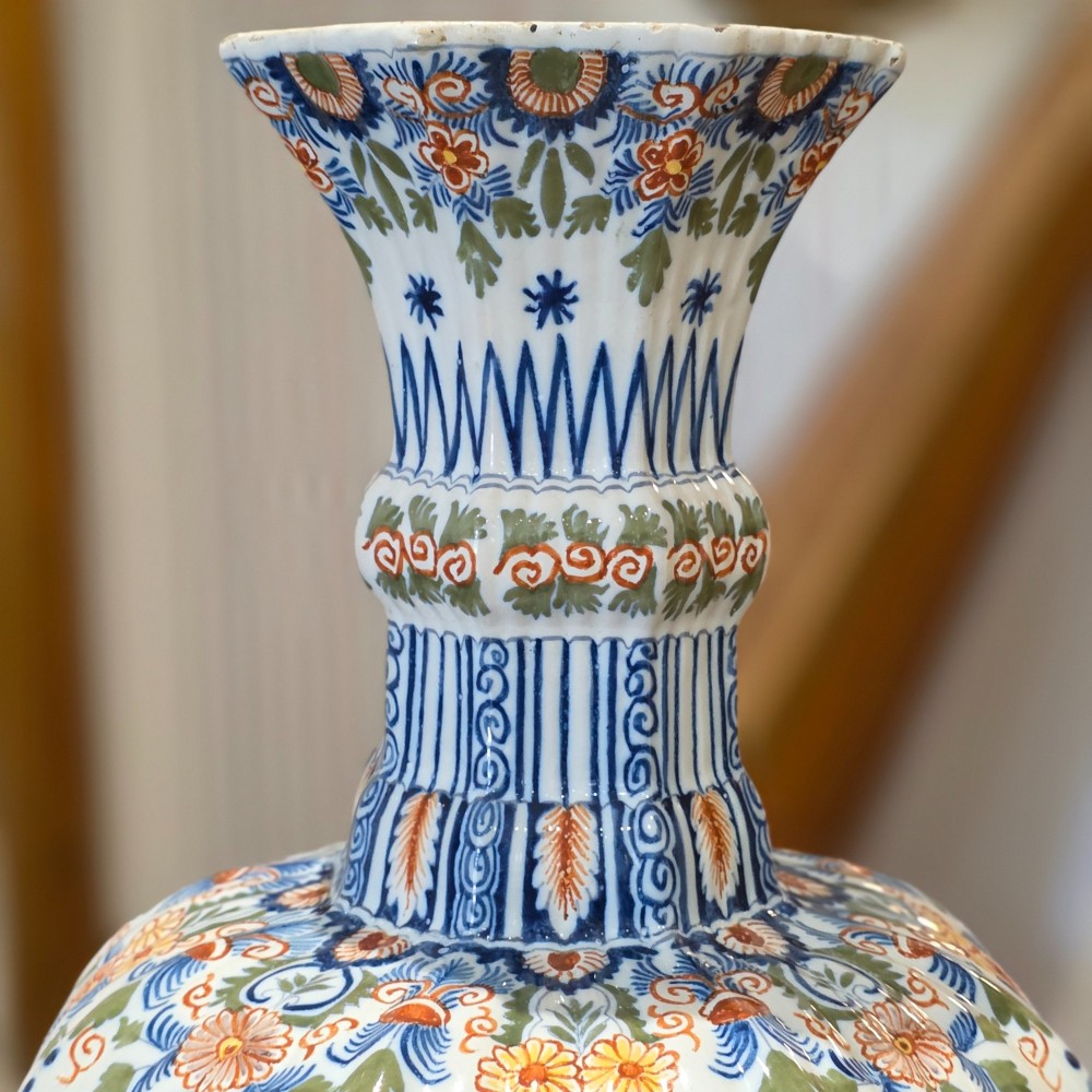 A tall nineteenth-century polychrome Delft earthenware vase featuring a fluted ribbed neck and bulbous body decorated with hand-painted floral motifs in blue, green, and orange on a white glazed ground. View VI