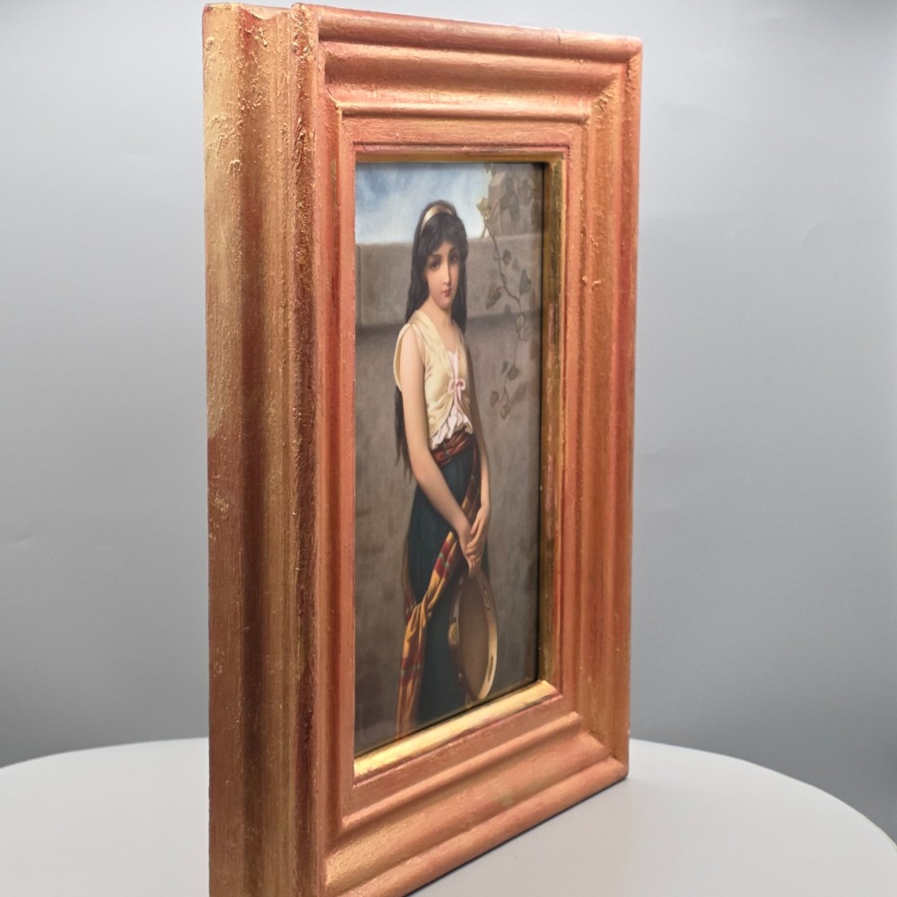 A rectangular hand-painted porcelain plaque depicting a young woman with a tambourine, set within a thick moulded gilt frame. View V