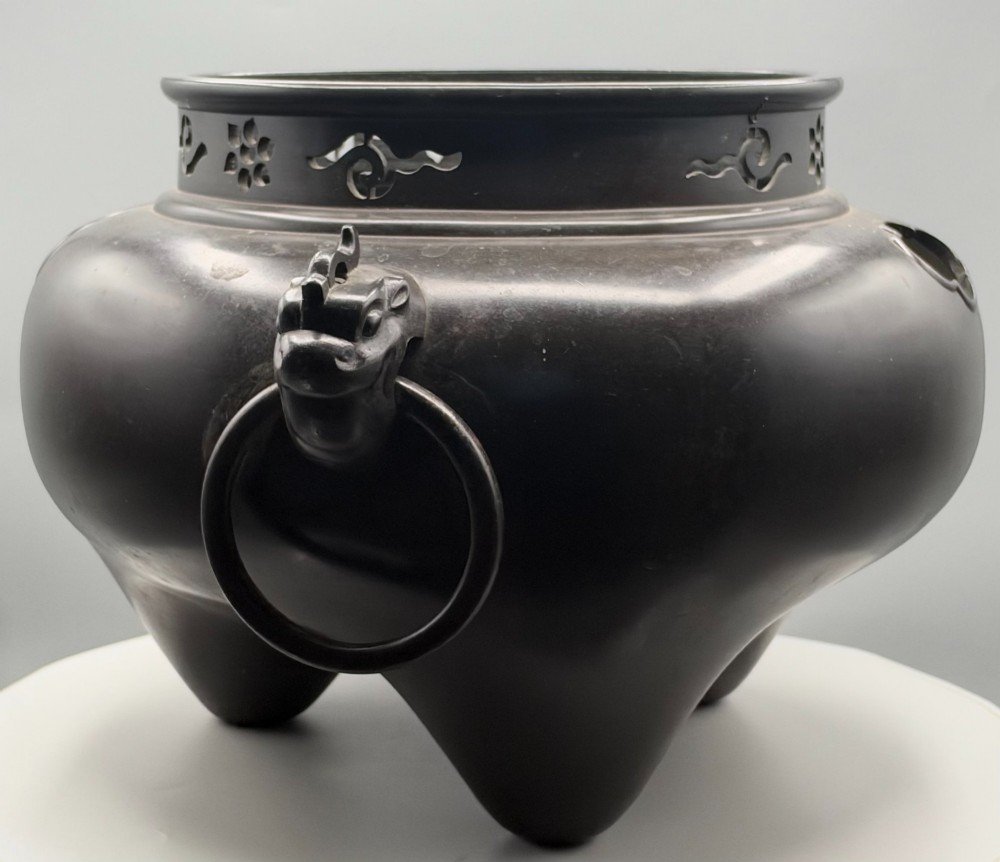 A dark patinated bronze tripod tea brazier featuring cast lion mask handles with loose ring pulls and pierced decorative motifs along the collar. View V