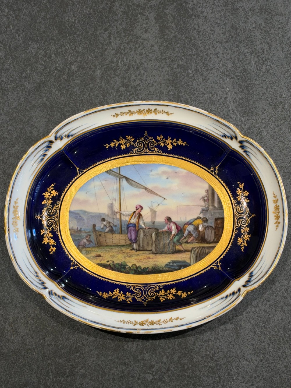 An oval French Sevres porcelain dish featuring a deep cobalt blue rim decorated with intricate gold leaf gilding surrounding a central hand-painted maritime harbour scene.