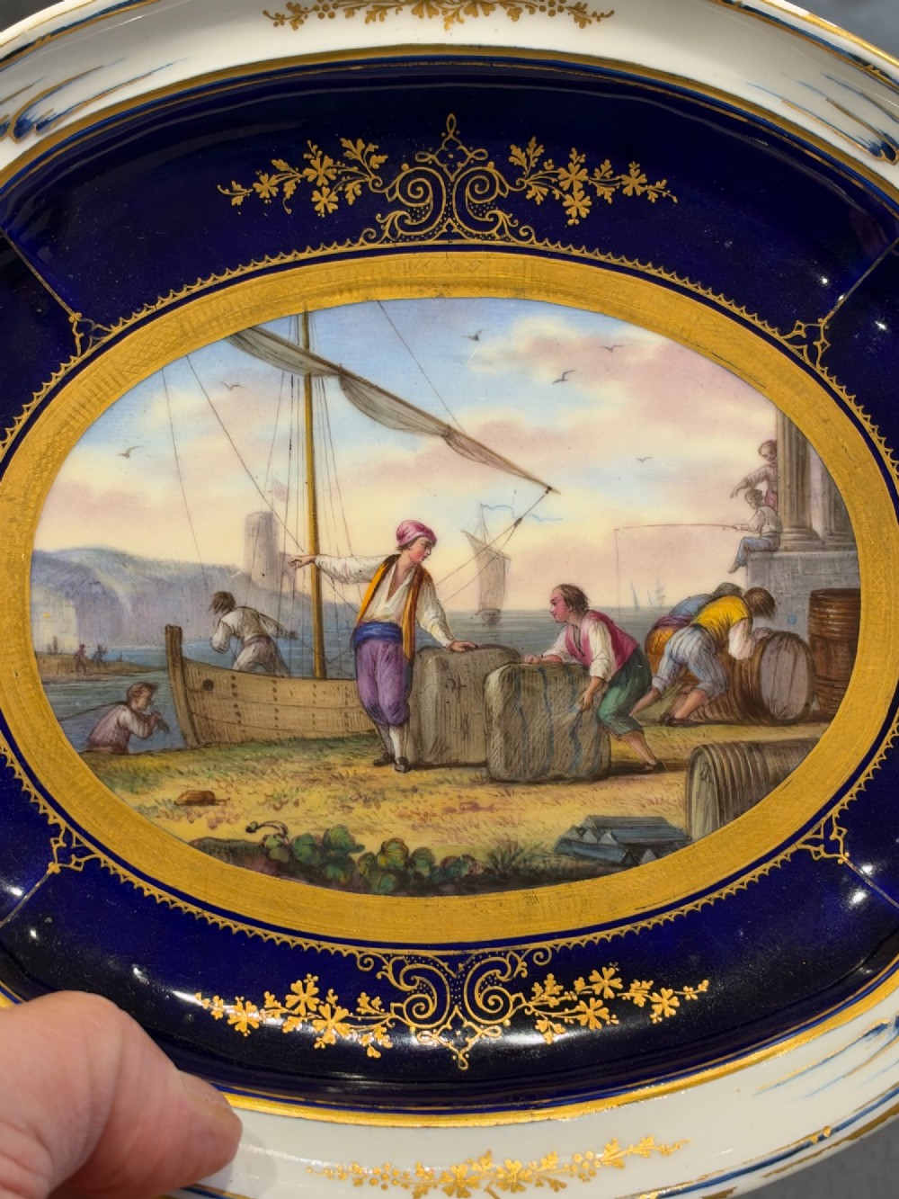 An oval French Sevres porcelain dish featuring a deep cobalt blue rim decorated with intricate gold leaf gilding surrounding a central hand-painted maritime harbour scene. View III