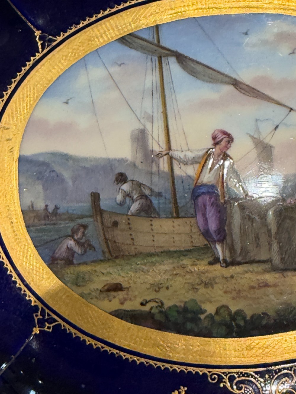 An oval French Sevres porcelain dish featuring a deep cobalt blue rim decorated with intricate gold leaf gilding surrounding a central hand-painted maritime harbour scene. View IV